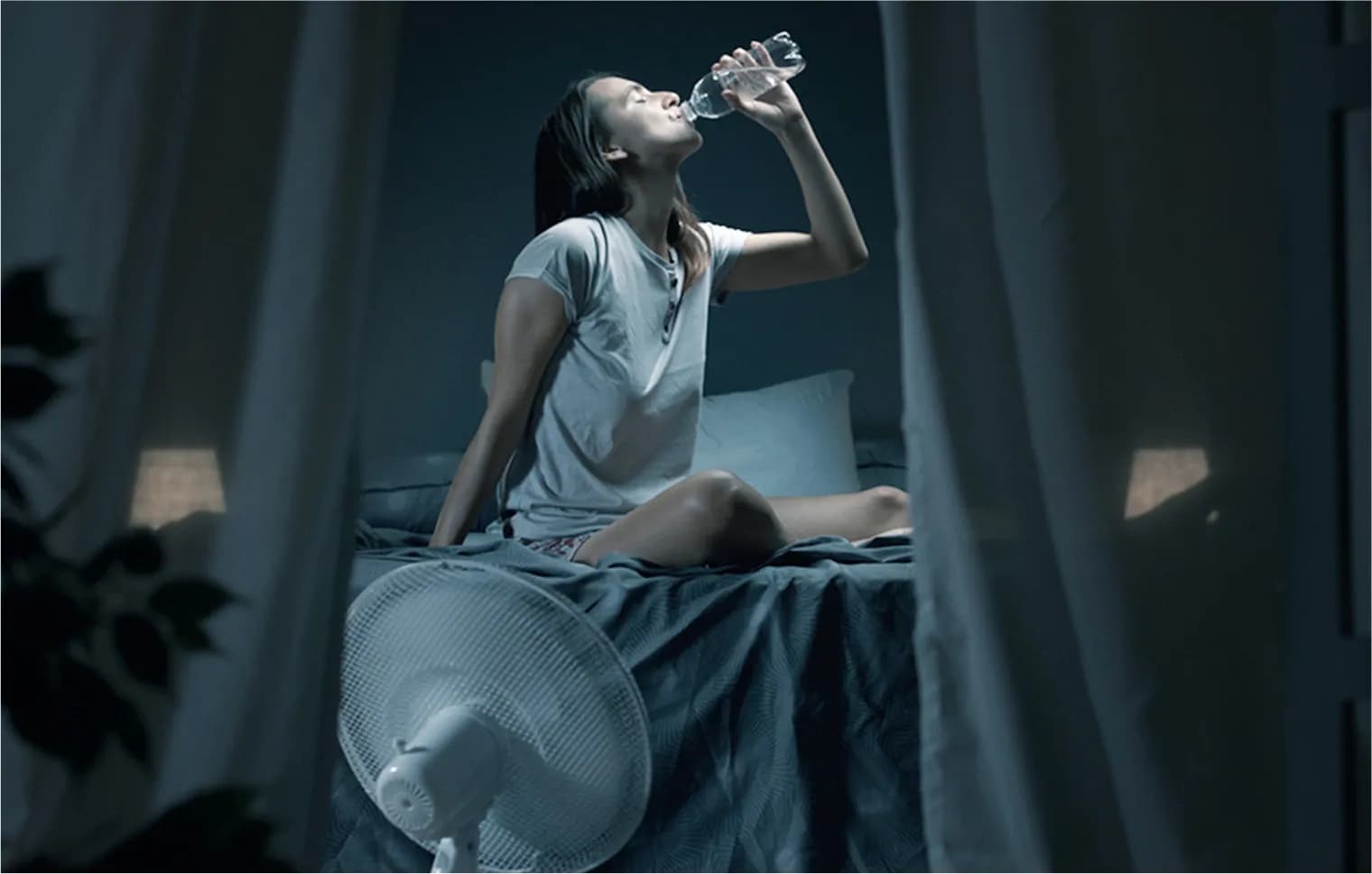A dehydrated woman chugging water on her bed in the middle of the night due to hot room temperature